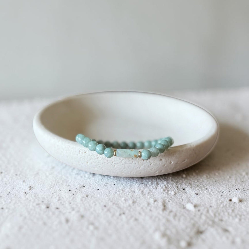 A light blue-green beaded bracelet with small gold spacer beads, resting inside a small white ceramic dish on a textured white surface.