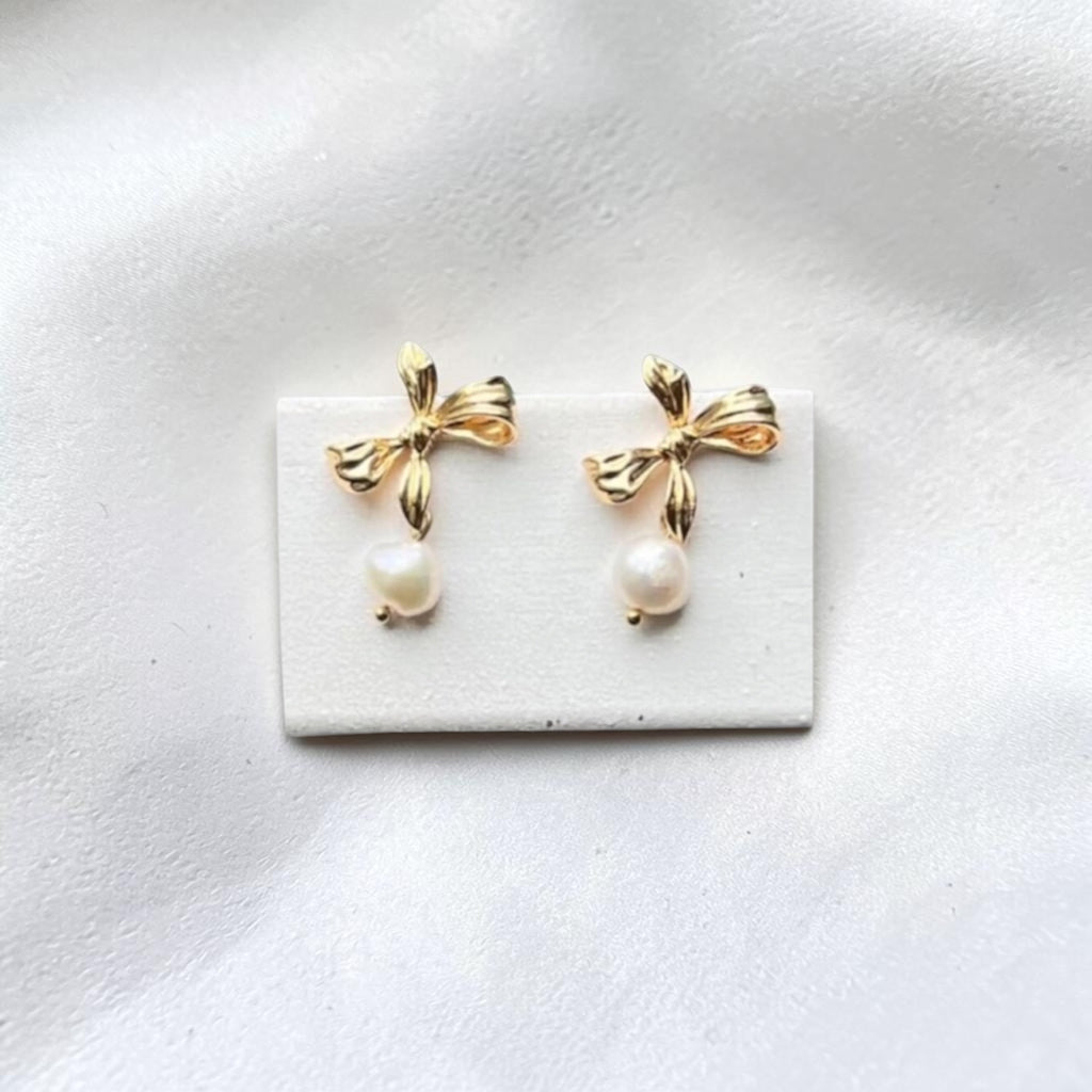 A pair of gold bow stud earrings with dangling white pearl drops, displayed on a small white rectangular card against a soft white fabric background.