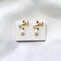 A pair of gold bow stud earrings with dangling white pearl drops, displayed on a small white rectangular card against a soft white fabric background.