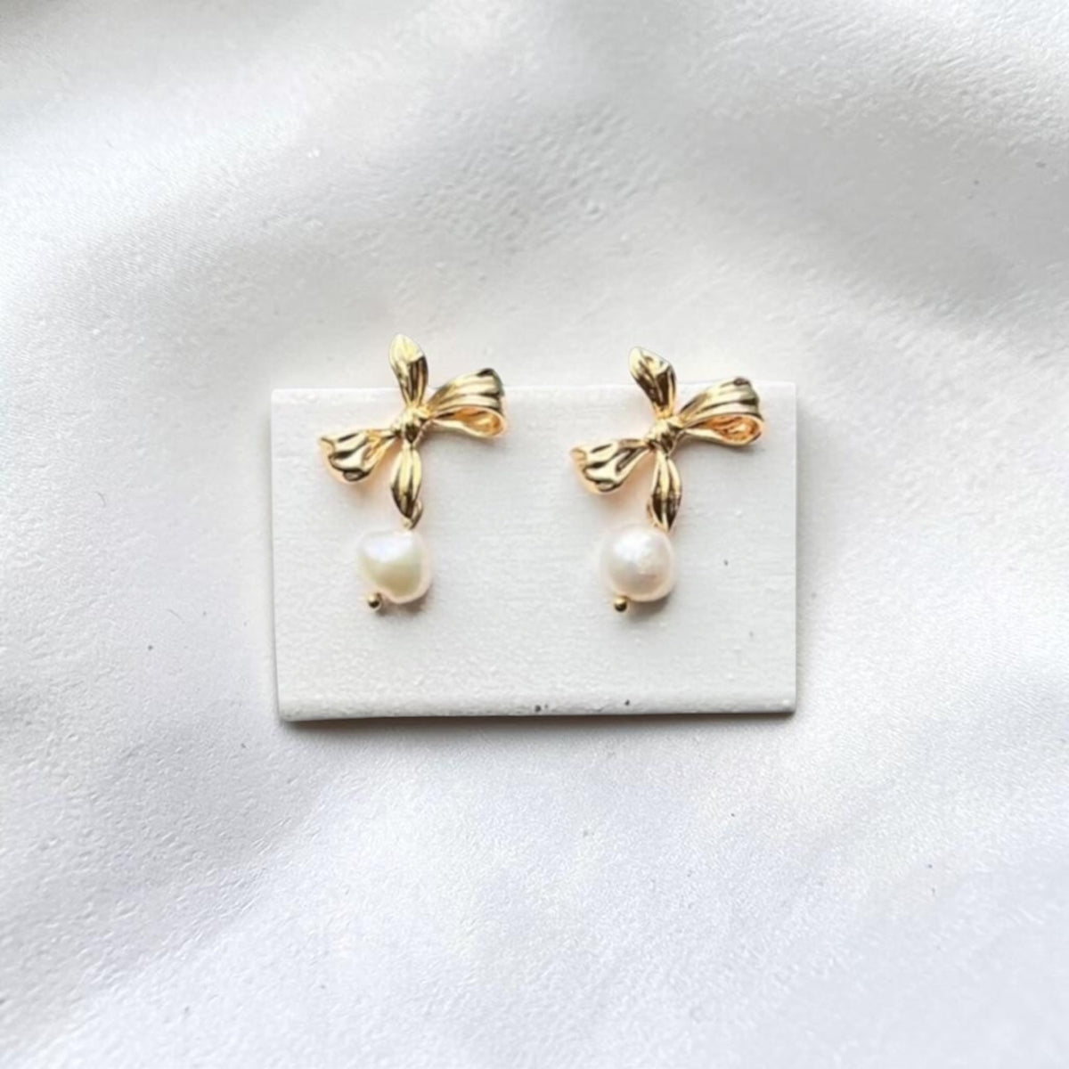 A pair of gold bow stud earrings with dangling white pearl drops, displayed on a small white rectangular card against a soft white fabric background.