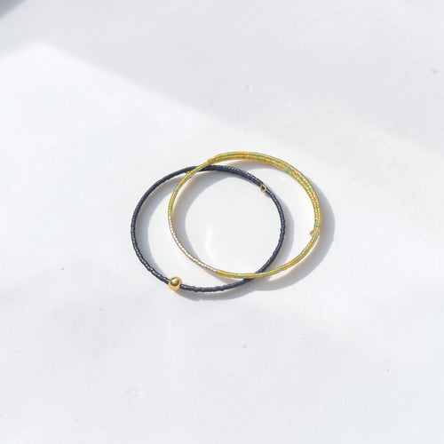 Two thin braided elastic bands, one in dark gray and one in textured gold, with a small gold bead accent on the gray band, displayed on a clean white surface.