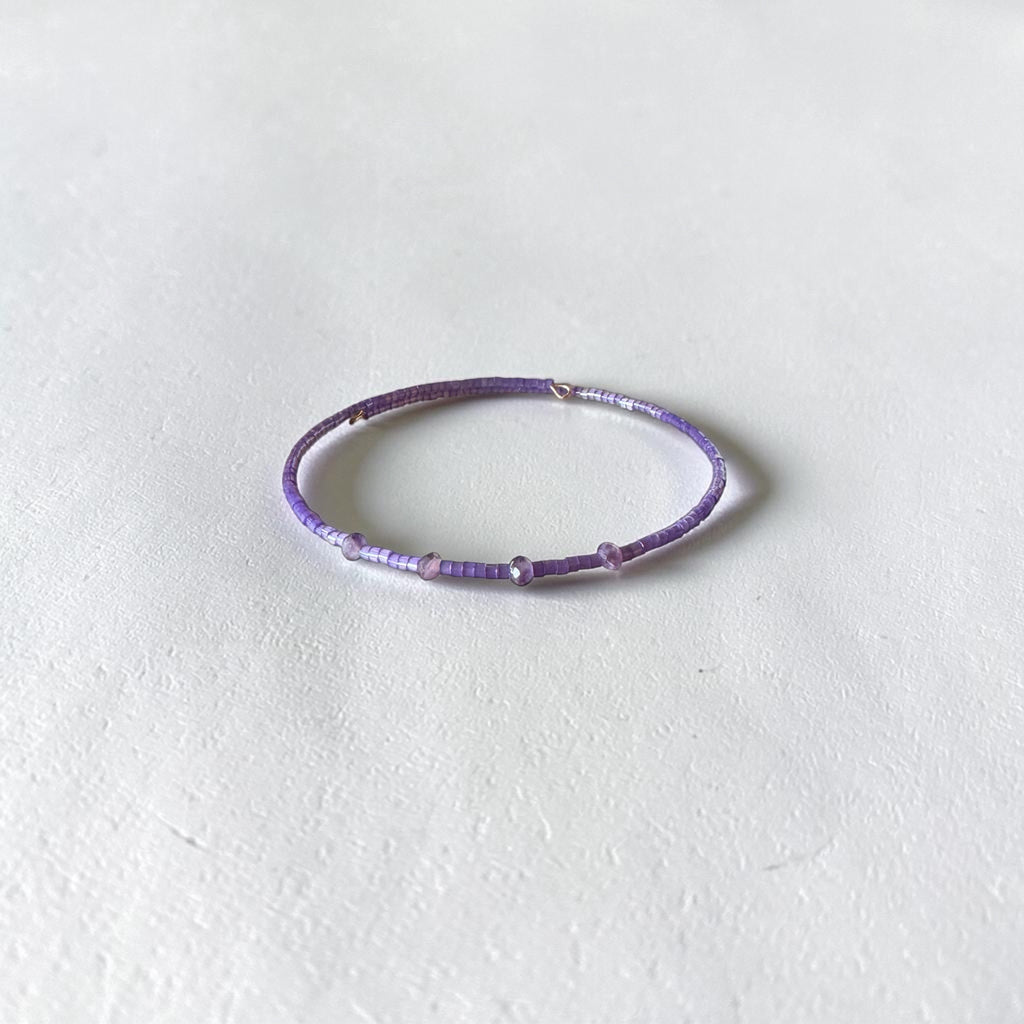 A thin beaded bracelet in shades of purple and lavender, featuring small metallic silver spacer beads, displayed on a textured off-white surface.