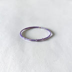 A thin beaded bracelet in shades of purple and lavender, featuring small metallic silver spacer beads, displayed on a textured off-white surface.