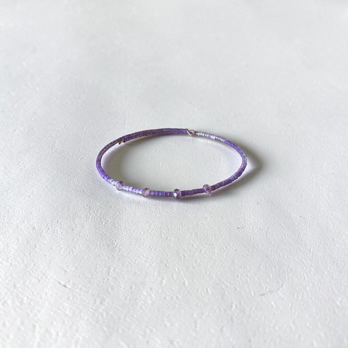 A thin beaded bracelet in shades of purple and lavender, featuring small metallic silver spacer beads, displayed on a textured off-white surface.