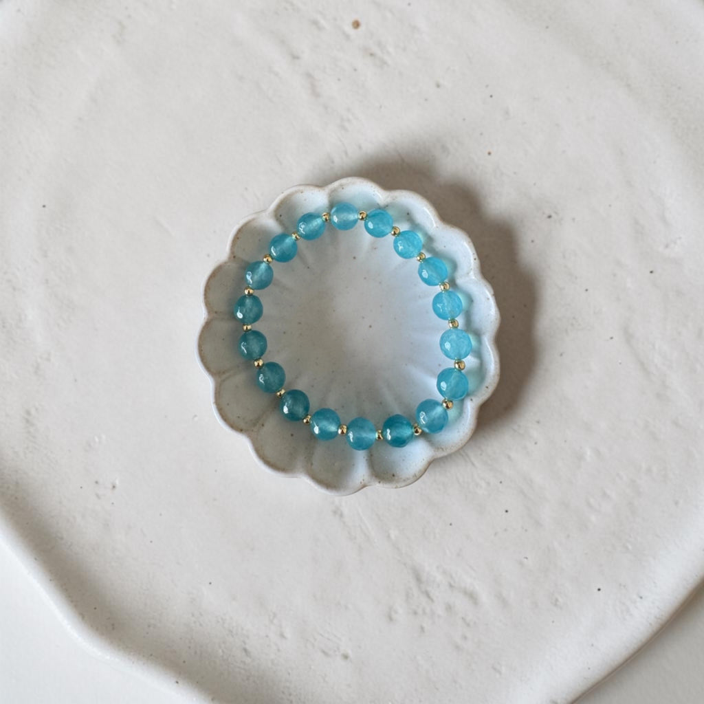 A beaded stretch bracelet made of light blue round beads separated by small gold spacer rings, displayed inside a small white scalloped dish on a textured off-white surface.