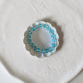A beaded stretch bracelet made of light blue round beads separated by small gold spacer rings, displayed inside a small white scalloped dish on a textured off-white surface.