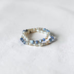 A delicate beaded bracelet with light blue and white beads, photographed on a textured white fabric background.