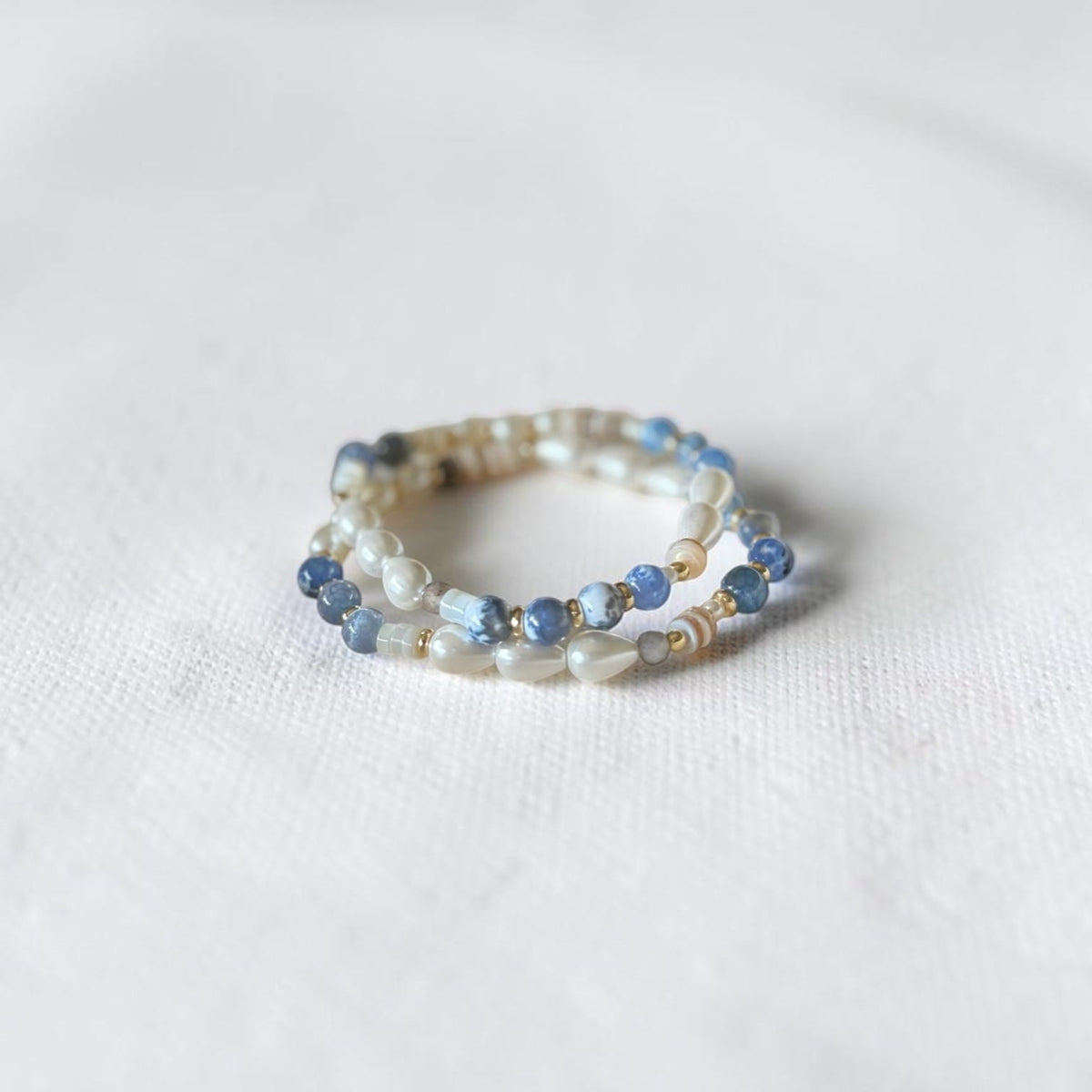 A delicate beaded bracelet with light blue and white beads, photographed on a textured white fabric background.