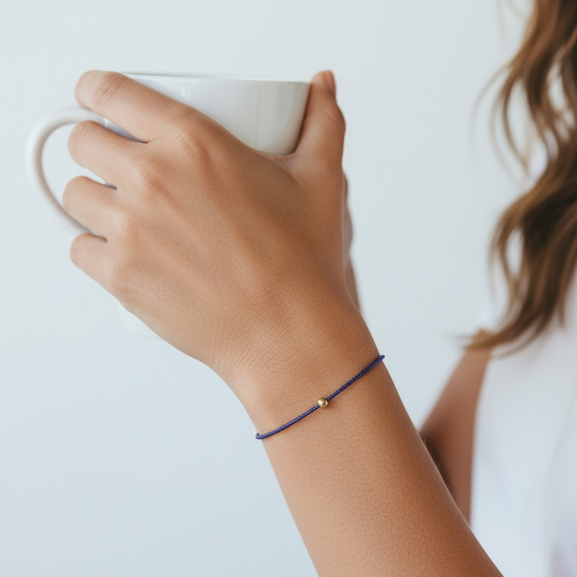 A close-up lifestyle shot of a person holding a white ceramic mug, showcasing a thin navy blue cord bracelet with a tiny gold bead on the wrist.