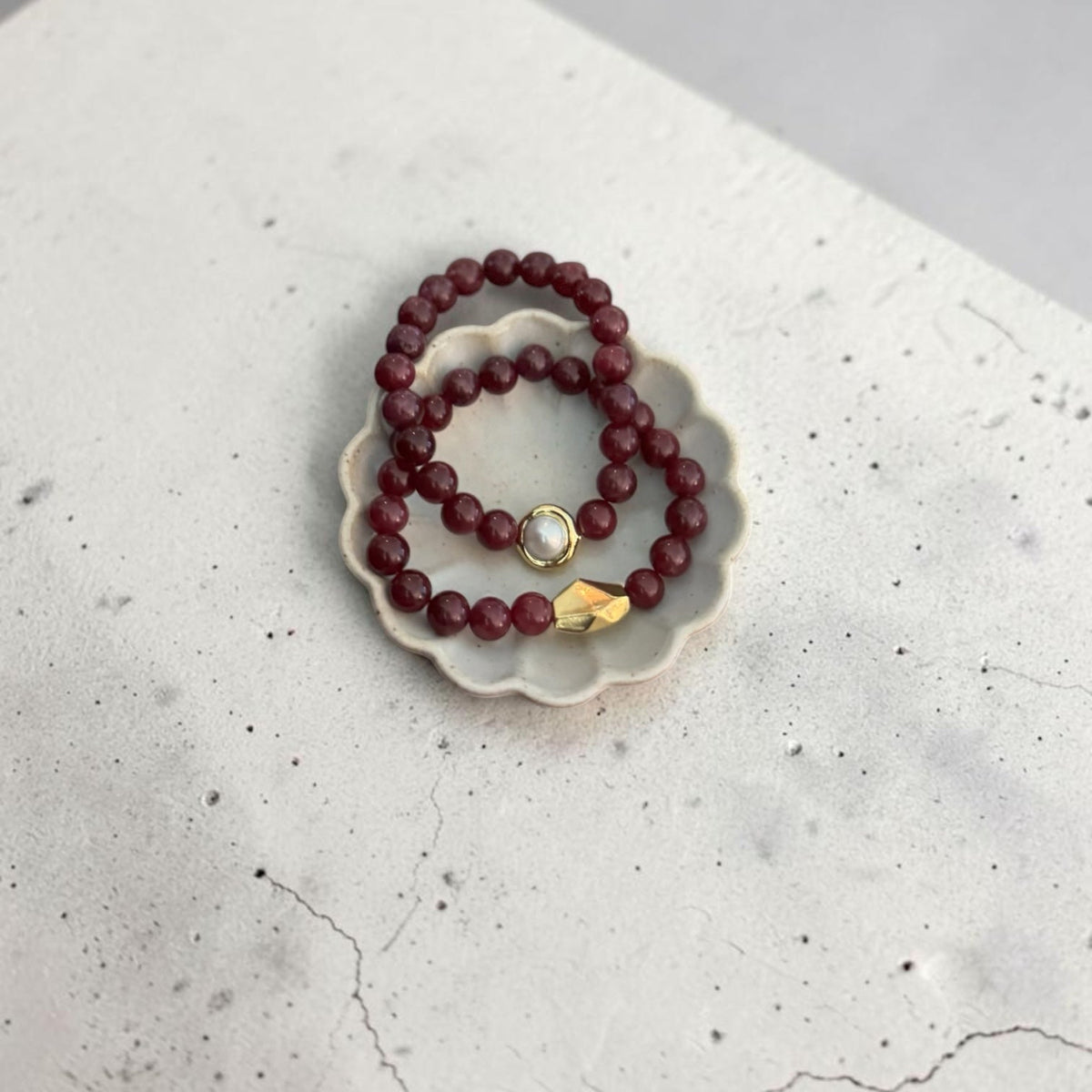 2 8mm Red Garnet Jade Bead Bracelet with a Gold And Pearl Accent and gold accent. Accent has a round pearl with gold surrounding the pearl itself. This bracelet is sitting in a dish.