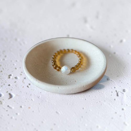 A beaded stretch bracelet with amber-yellow beads and a single white pearl-like bead, displayed inside a small round ceramic dish on a textured light gray surface.