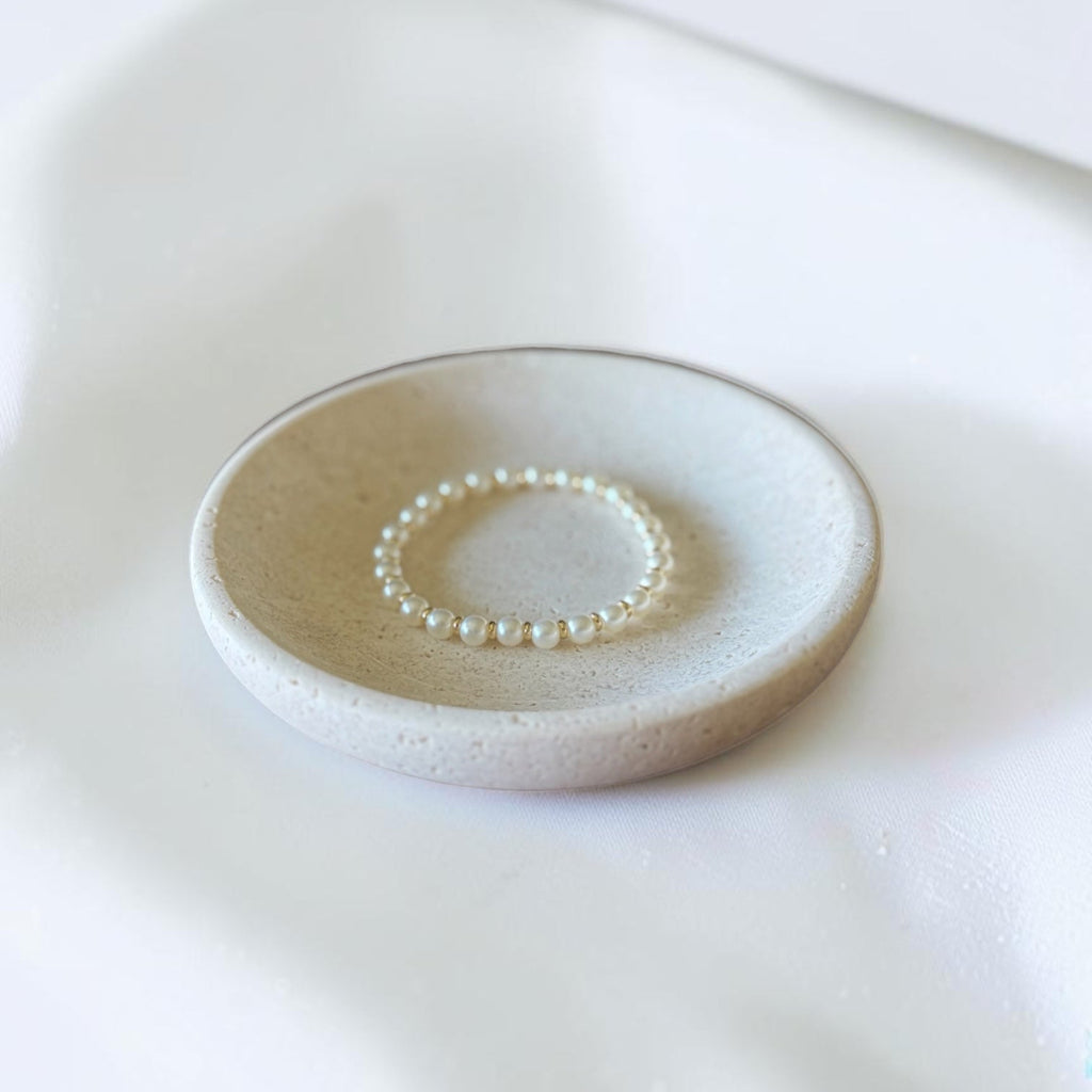 A delicate gold beaded bracelets displayed on a small, round, textured ceramic dish over a soft white fabric background.