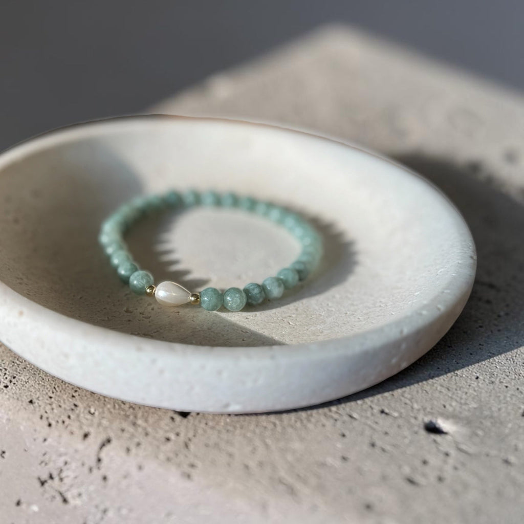 A beaded stretch bracelet with light green beads and a white teardrop pearl charm, displayed on a small white ceramic dish over a textured concrete surface.