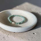 A beaded stretch bracelet with light green beads and a white teardrop pearl charm, displayed on a small white ceramic dish over a textured concrete surface.