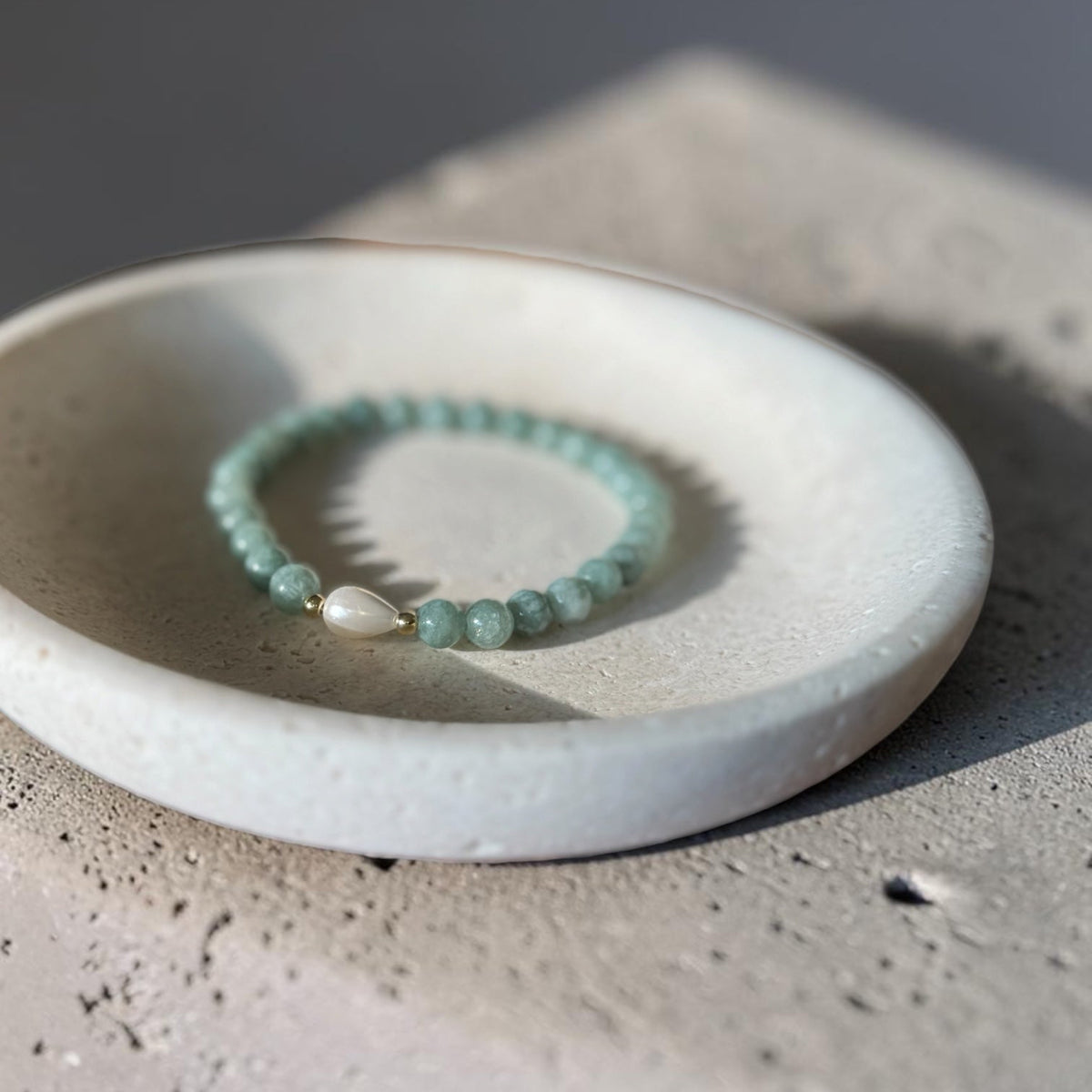 A beaded stretch bracelet with light green beads and a white teardrop pearl charm, displayed on a small white ceramic dish over a textured concrete surface.