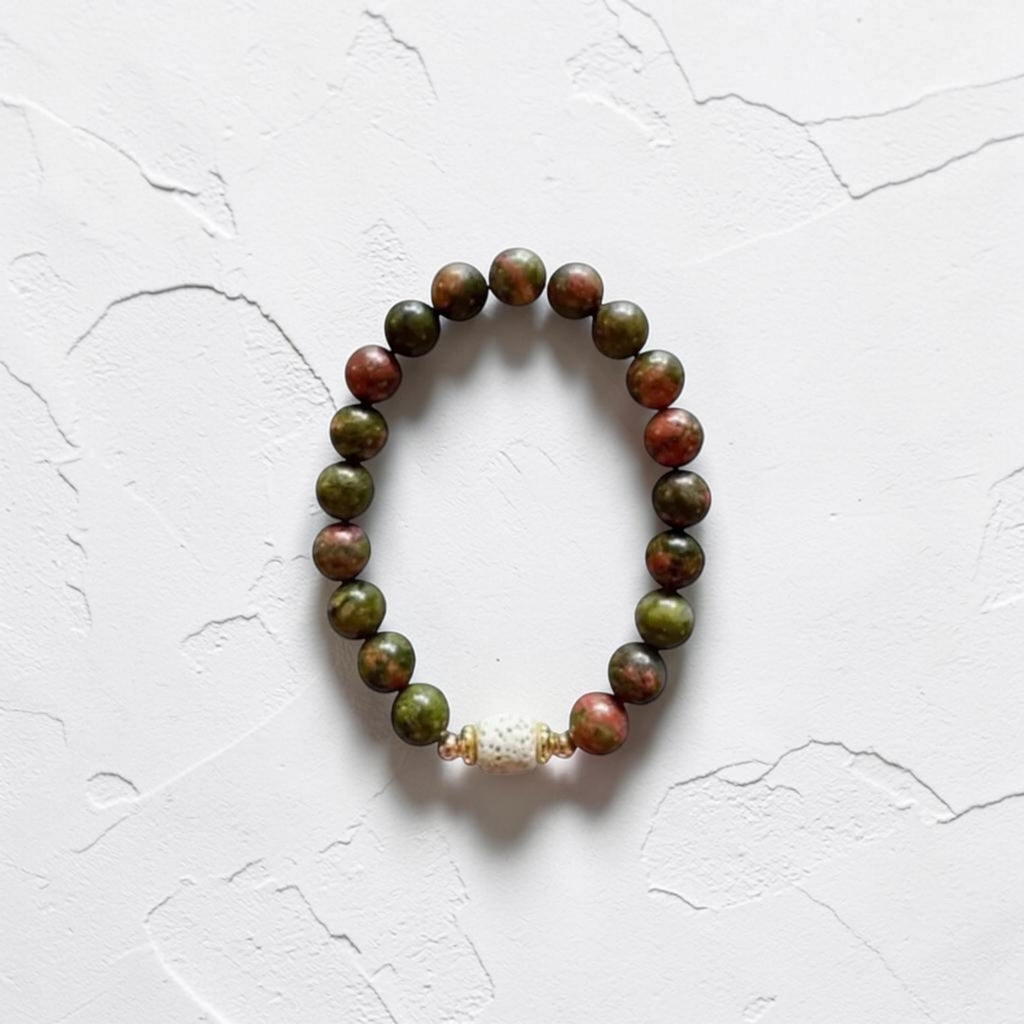 A beaded stretch bracelet made of round green and reddish-brown beads, with a small white textured accent bead near the bottom, displayed on a white textured background.