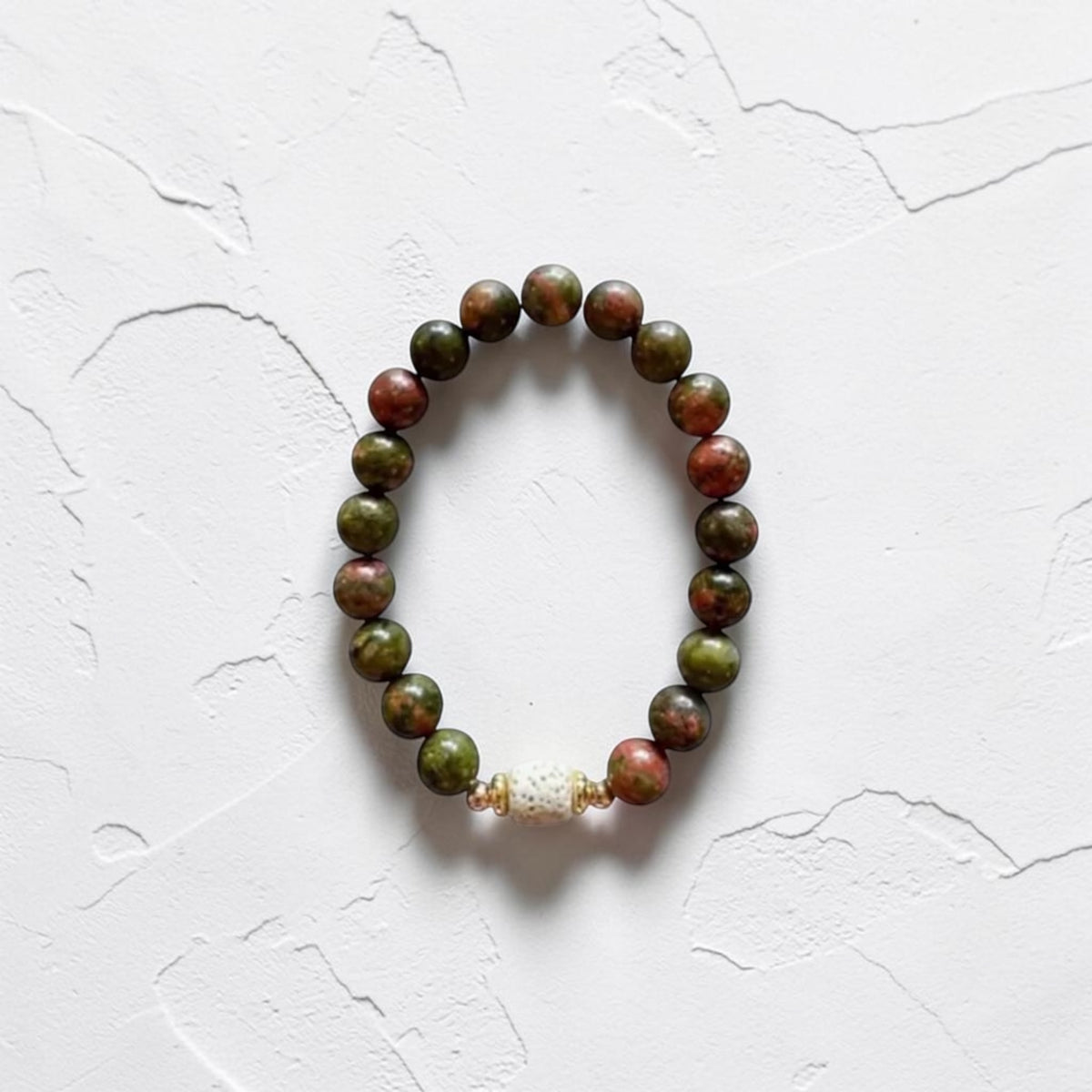 A beaded stretch bracelet made of round green and reddish-brown beads, with a small white textured accent bead near the bottom, displayed on a white textured background.