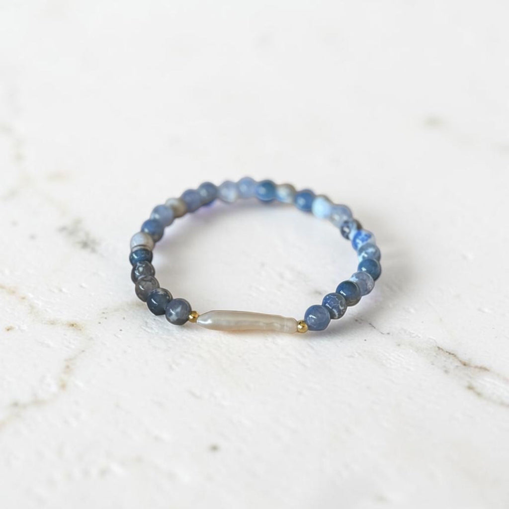 A beaded stretch ring featuring round blue and gray beads with a central translucent white teardrop bead and small gold spacer accents, resting on a light marble surface.
