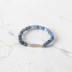A beaded stretch ring featuring round blue and gray beads with a central translucent white teardrop bead and small gold spacer accents, resting on a light marble surface.