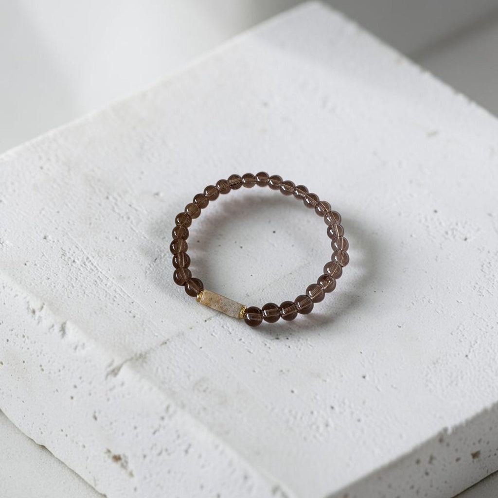 A beaded stretch bracelet with dark brown translucent beads and a small gold-toned rectangular accent, displayed on a textured white stone block.