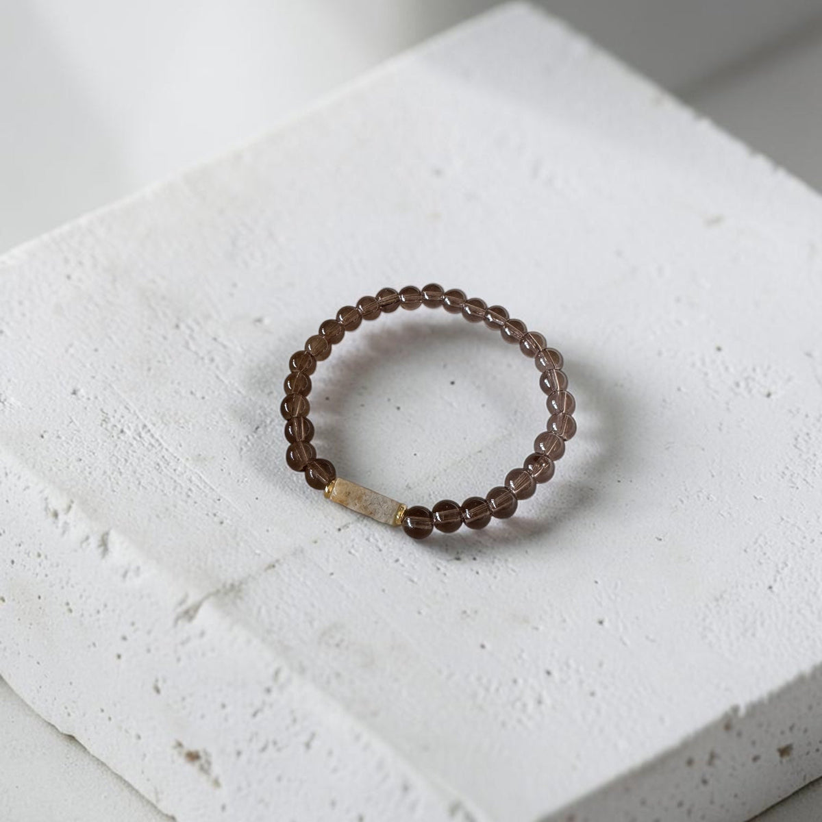 A beaded stretch bracelet with dark brown translucent beads and a small gold-toned rectangular accent, displayed on a textured white stone block.