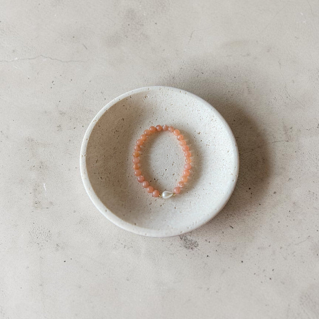 A peach-orange beaded bracelet with a small white bead accent, displayed inside a small speckled white ceramic dish on a textured light gray surface.