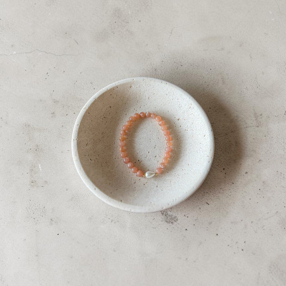 A peach-orange beaded bracelet with a small white bead accent, displayed inside a small speckled white ceramic dish on a textured light gray surface.