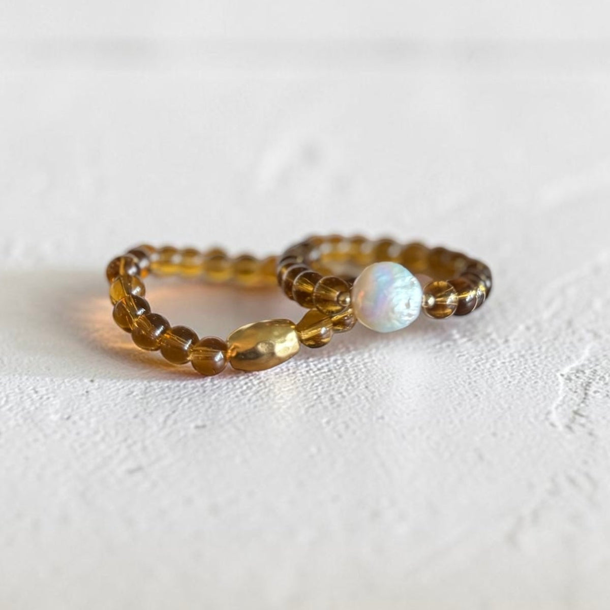 Two beaded bracelets on a textured white surface. One ring features translucent amber-brown beads, and the other features translucent amber-brown beads with a single white iridescent opal bead.