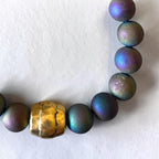 A close-up of a beaded strand featuring round iridescent beads that shift between green, blue, and purple, accented by a large textured gold cylindrical bead in the center, laid on a white surface.