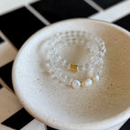 A stack of two beaded bracelets, including clear faceted beads, white pearl beads, and a gold clasp, displayed on a small speckled cream ceramic dish over a black and white checkered tile surface.