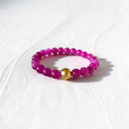 A beaded stretch bracelet featuring faceted magenta-purple beads and a single polished gold accent bead, displayed on a textured white surface.