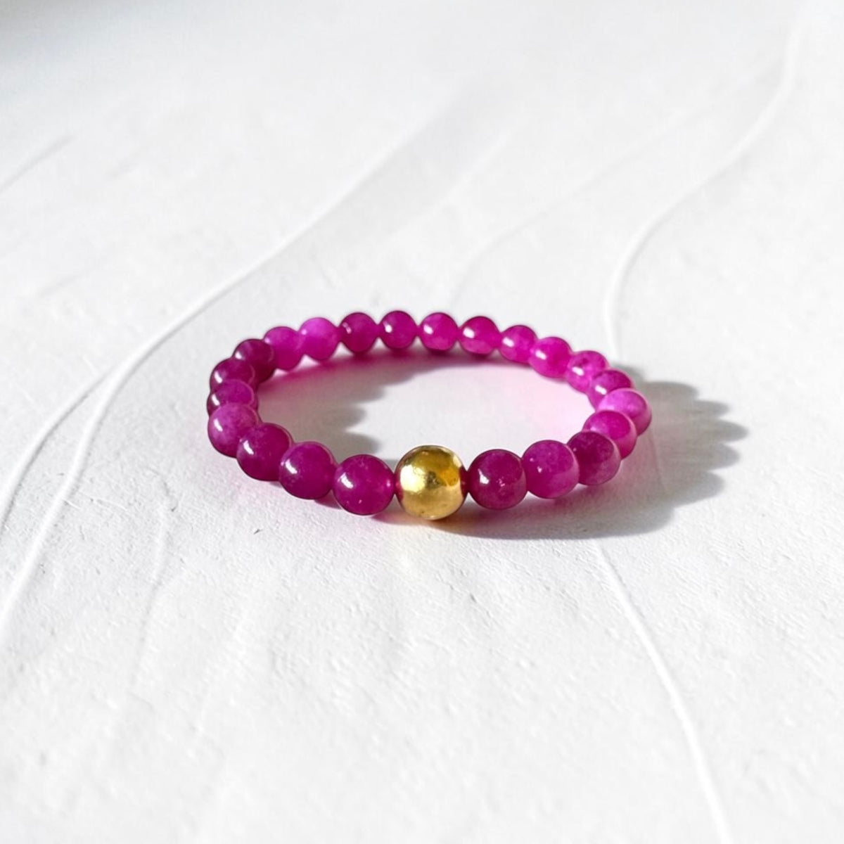 A beaded stretch bracelet featuring faceted magenta-purple beads and a single polished gold accent bead, displayed on a textured white surface.