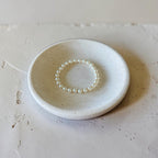 A delicate pearl beaded bracelet displayed inside a small, round, textured off-white ceramic dish on a matching neutral surface.