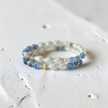 A beaded stretch bracelet featuring white iridescent teardrop beads, light blue faceted beads, and small gold spacer rings, resting on a textured white surface.