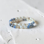 A beaded stretch bracelet featuring white iridescent teardrop beads, light blue faceted beads, and small gold spacer rings, resting on a textured white surface.