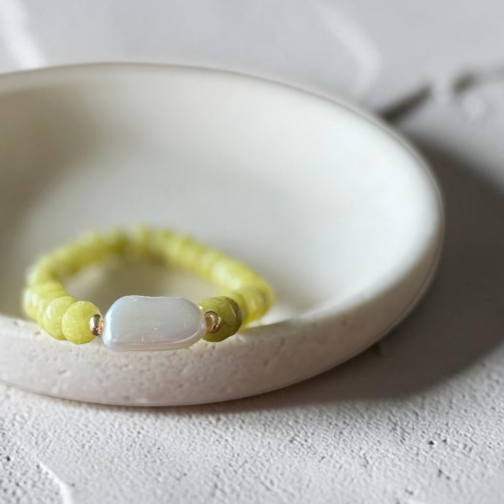 A beaded stretch bracelet with pale yellow round beads, small metallic spacer beads, and a central white stone, displayed on a textured white ceramic dish.