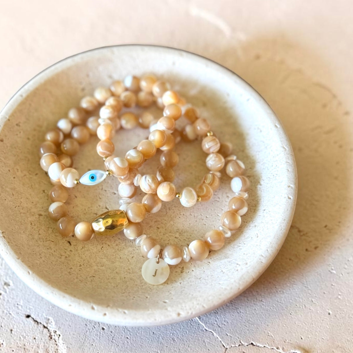 A set of beaded bracelets with light beige and white pearls, a small blue evil eye bead, and a gold accent, displayed in a small speckled ceramic dish.