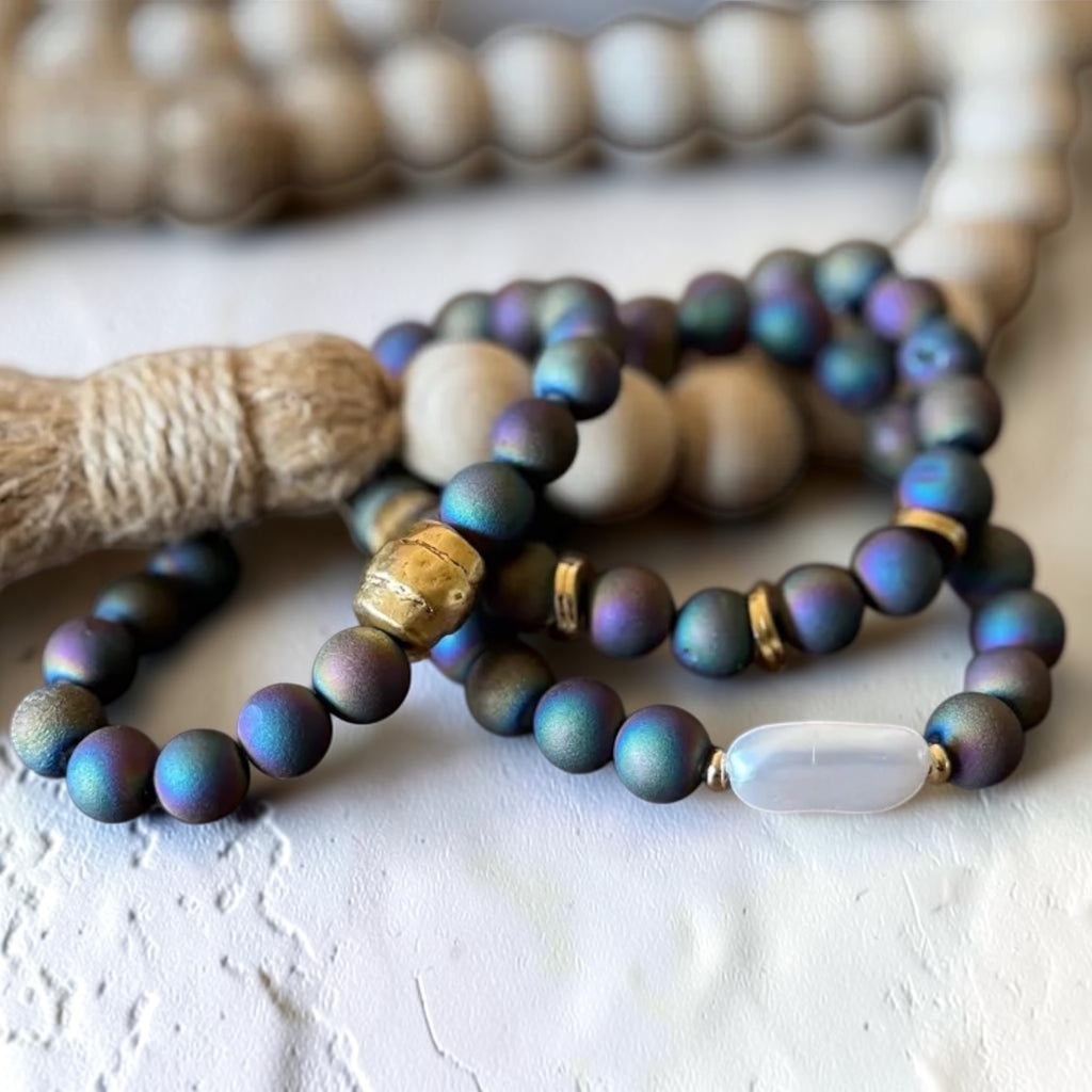 A close-up of a beaded strand bracelet with iridescent dark blue and purple beads, accented by a gold Buddha bead, a white translucent bead, and light wood beads in the background.