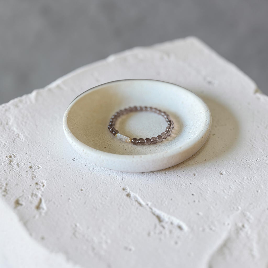 A beaded stretch ring with translucent brown and white beads rests inside a small, round, off-white ceramic dish on a textured white stone slab against a neutral gray background.