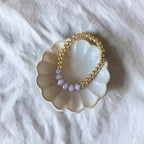 A gold beaded stretch bracelet with a row of small lavender beads, displayed on a scalloped cream ceramic dish over a white fabric background.
