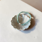 A light green beaded bracelet with a small clear teardrop charm, displayed on a white ceramic flower-shaped dish against a warm beige background.
