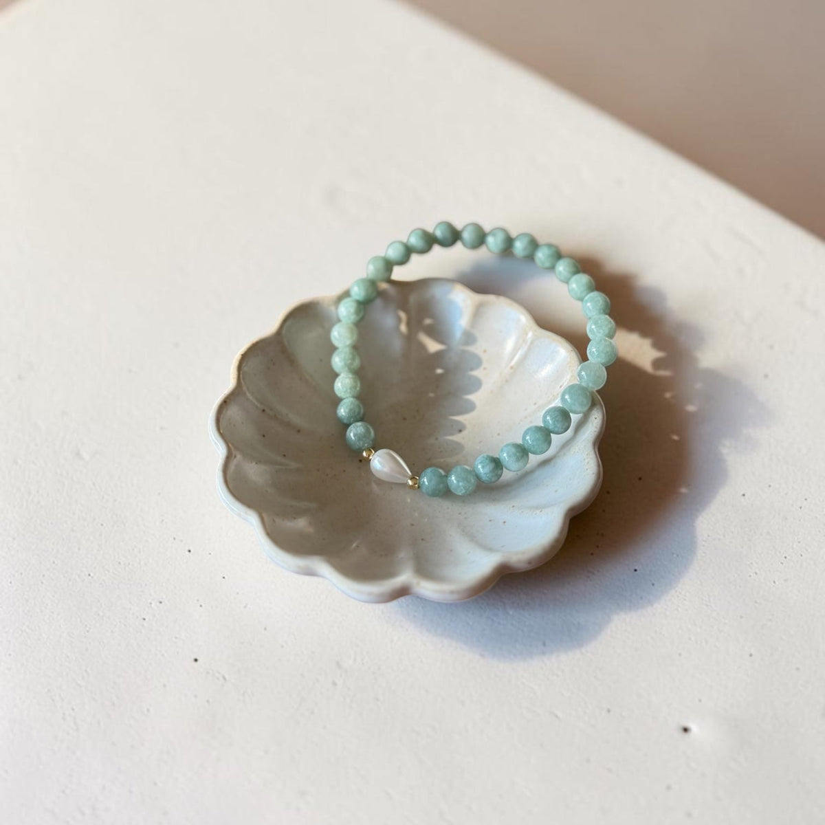 A light green beaded bracelet with a small clear teardrop charm, displayed on a white ceramic flower-shaped dish against a warm beige background.