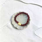 A beaded stretch bracelet with deep red beads and a small gold-toned geometric charm, displayed on a scalloped white dish over a white surface.