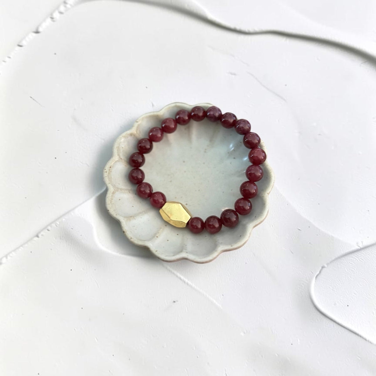 A beaded stretch bracelet with deep red beads and a small gold-toned geometric charm, displayed on a scalloped white dish over a white surface.