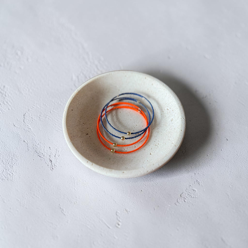 A set of thin wire bracelets in blue, orange, and gold, displayed inside a small speckled ceramic dish on a textured light gray surface.