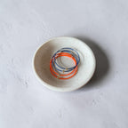 A set of thin wire bracelets in blue, orange, and gold, displayed inside a small speckled ceramic dish on a textured light gray surface.