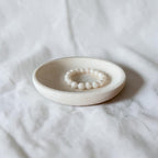 A cream beaded bracelet displayed inside a shallow round ceramic dish on a soft white fabric background.