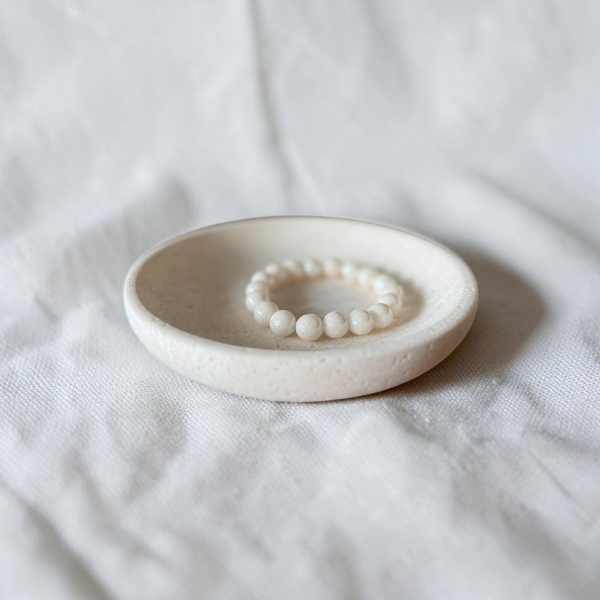 A cream beaded bracelet displayed inside a shallow round ceramic dish on a soft white fabric background.