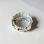 A stack of delicate beaded stretch bracelets in white, light blue, and beige, displayed on a small scalloped white dish against a neutral off-white background.