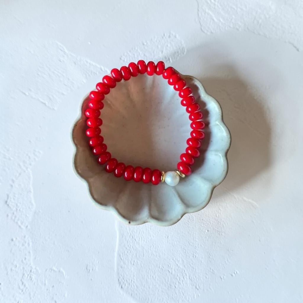 A red beaded bracelet with a small white pearl accent, displayed on a scalloped ceramic dish against a textured white background.